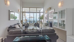 Un salon moderne avec une table basse en verre, des canapés gris et une vue sur l’océan grâce à de grandes fenêtres.