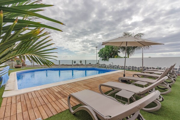 Une piscine dotée de chaises longues et d’un parasol, entourée de verdure et offrant une vue sur la mer.