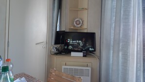 A room with a television on a wooden stand, a small shelf with a decorative item, and a curtain partially drawn. - Capri 39 (Sigean)