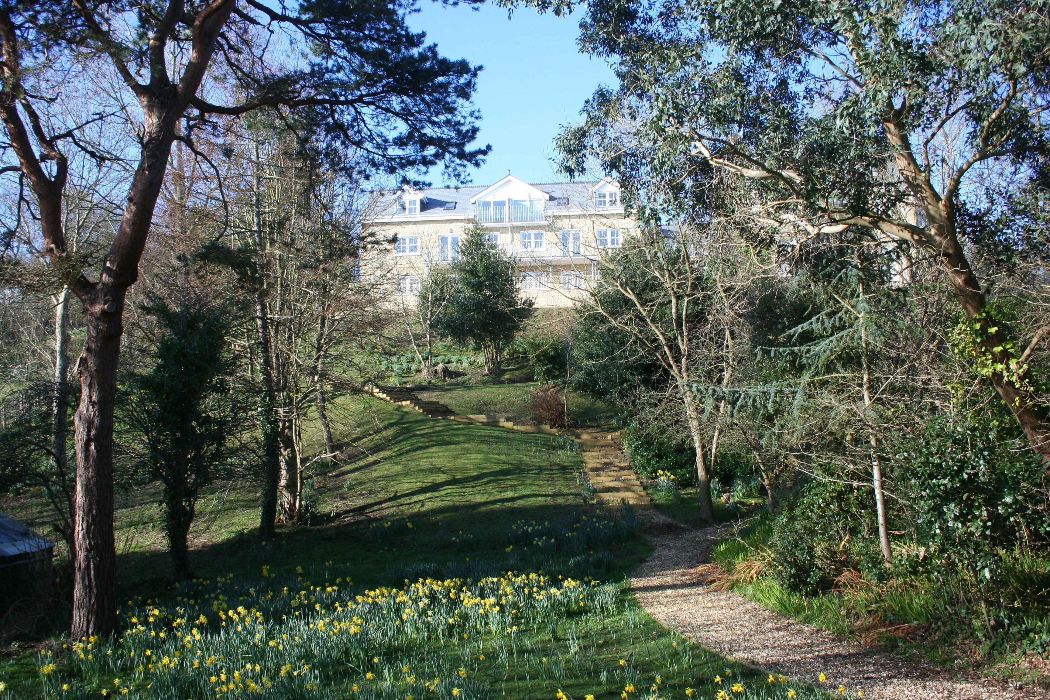 A large, multi-story building with a symmetrical facade, surrounded by a well-maintained garden with a variety of trees and a grassy area with yellow flowers.