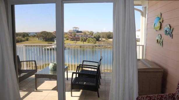 A balcony with a view of a lake and a house across the water. There are two chairs and a table on the balcony. - Villa Sabine by Surfside VR (Pensacola Beach)