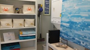 A room with a white shelf holding several bags with crab designs, a vacuum cleaner, a washing machine, and a blue wall with a wave pattern. - Villa Sabine by Surfside VR (Pensacola Beach)