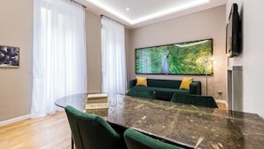 A modern living room with a curved dark marble table, green upholstered chairs, a green sofa with yellow cushions, and a large peacock feather painting on the wall.