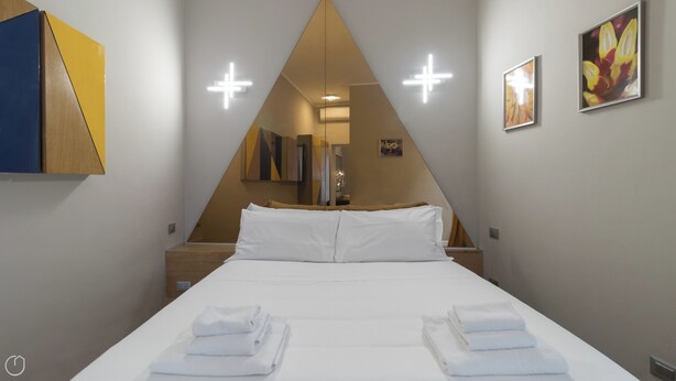 A hotel room with a bed, white linens, a mirror, and a painting on the wall.