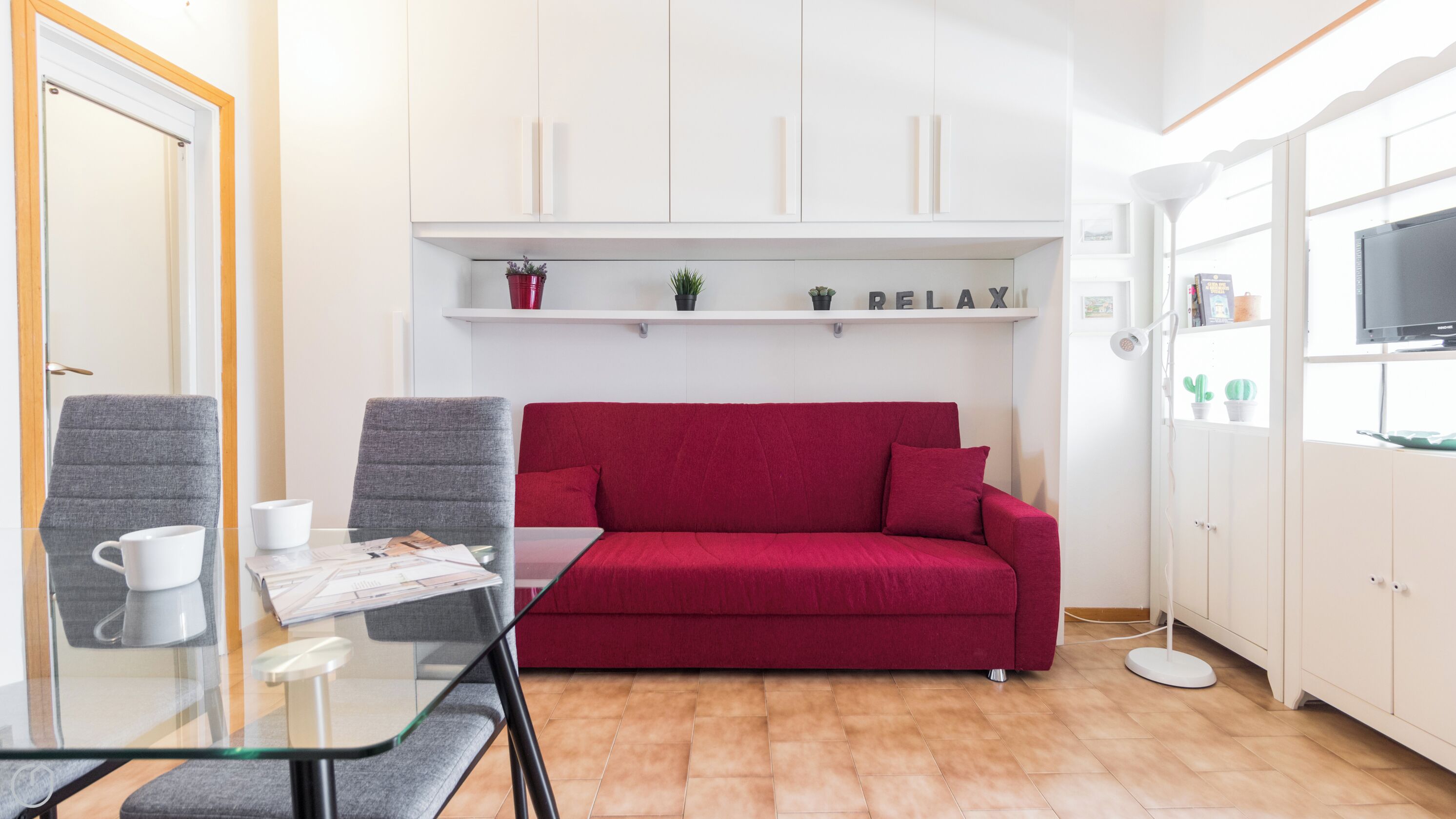 Un espace de vie compact comprenant une table en verre, deux chaises, un canapé rouge, un meuble blanc et un téléviseur placé à droite.
