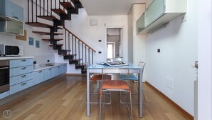 A modern kitchen with a wooden staircase, a dining area with a glass table and metal chairs, and a visible microwave. - Italianway Easy \- Baiamonti 1 (Milan)