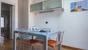 A dining area with a glass table, metal chairs, and a painting of a lighthouse on the wall. - Italianway Easy \- Baiamonti 1 (Milan)