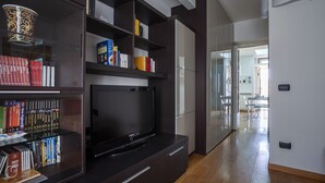 A modern living room with a flat-screen TV, built-in shelving with books, and a dining area with a glass partition. - Italianway Easy \- Baiamonti 1 (Milan)