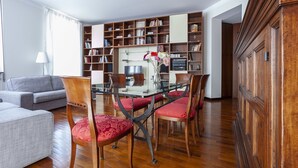 A living room with a glass-top dining table, red upholstered chairs, a grey sofa, and a wooden bookshelf filled with books. - Italianway - Lipari 8 (Milan)