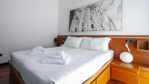 A neatly made bed with white linens and towels, a wooden headboard, a nightstand with a white lamp, and a framed picture of a cathedral on the wall. - Italianway - Lipari 8 (Milan)