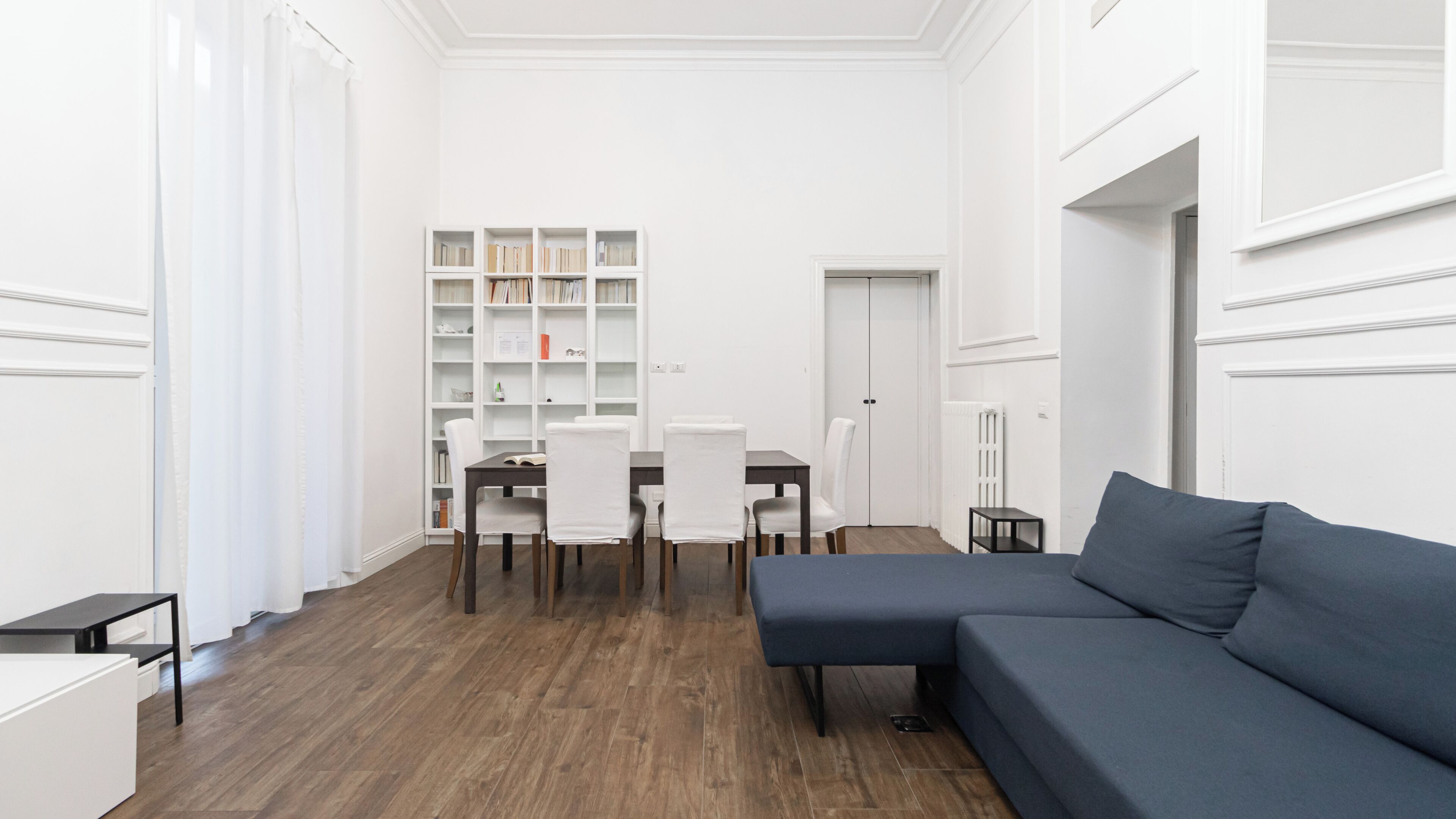 A modern living room with a dark blue sectional sofa, a dining table with chairs, and a white bookshelf.