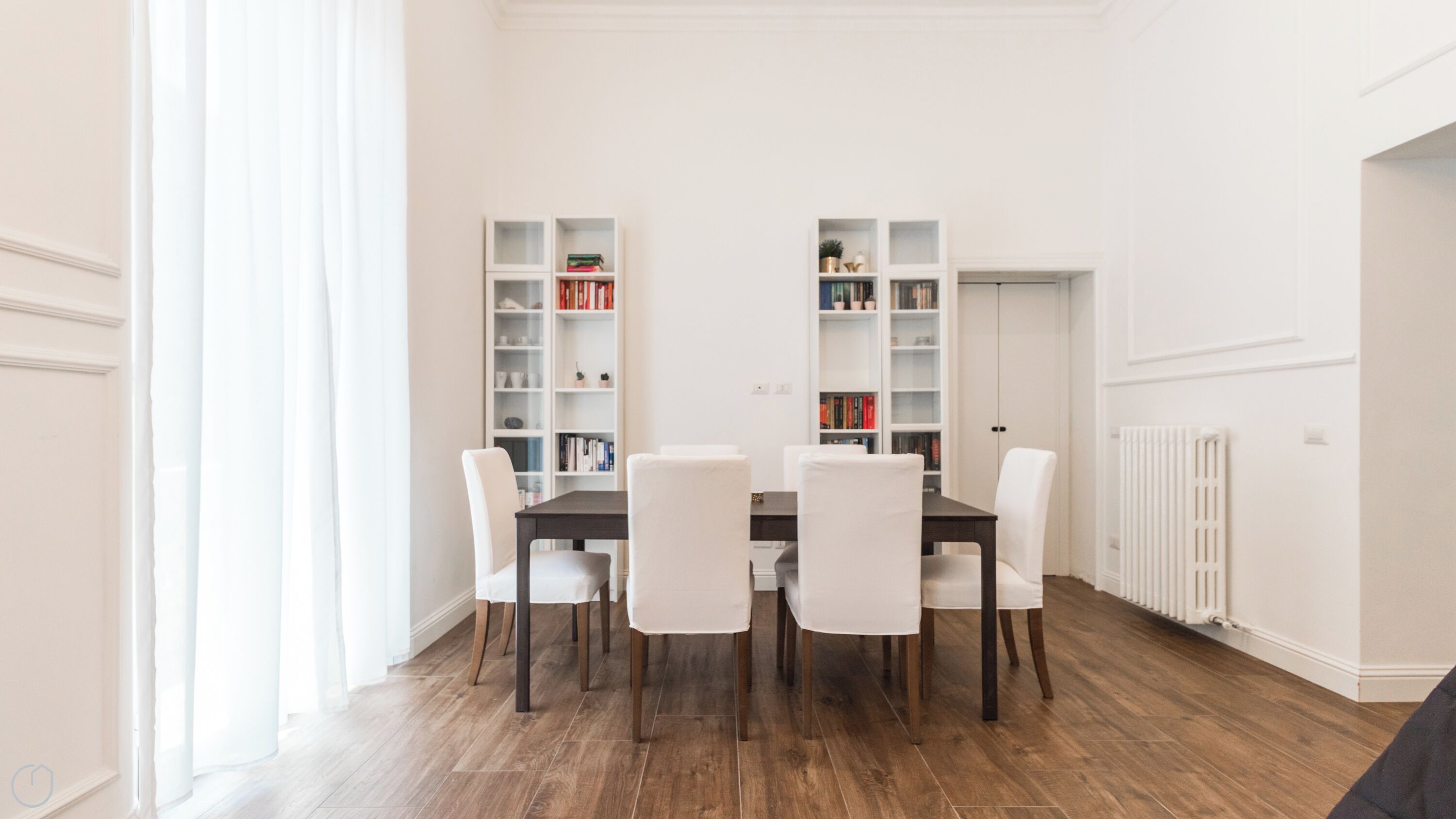 Un comedor moderno con una mesa de madera oscura y sillas blancas, flanqueado por estanterías blancas llenas de libros y objetos decorativos.
