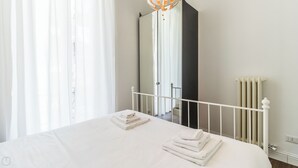 Una cama bien tendida con sábanas y toallas blancas, cabecera de metal blanco, un radiador y un espejo grande que refleja la habitación.