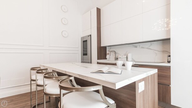 Una cocina moderna con encimera de mármol, armarios blancos y horno empotrado. Hay tres sillas blancas con estructura metálica y cojines blancos.