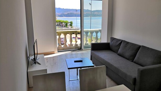 A modern living room with a grey sofa, a small table, and a chair. A large sliding glass door leads to a balcony with a view of the sea and a distant mountain range. - Superior Sea View Apartment (APT 2) (Poio)