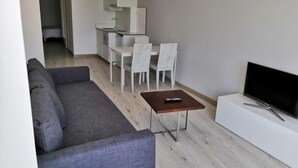 A modern living room with a grey sofa, a small wooden coffee table, a TV on a white cabinet, and a dining area with white chairs and a small kitchen in the background. - Superior Sea View Apartment (APT 2) (Poio)