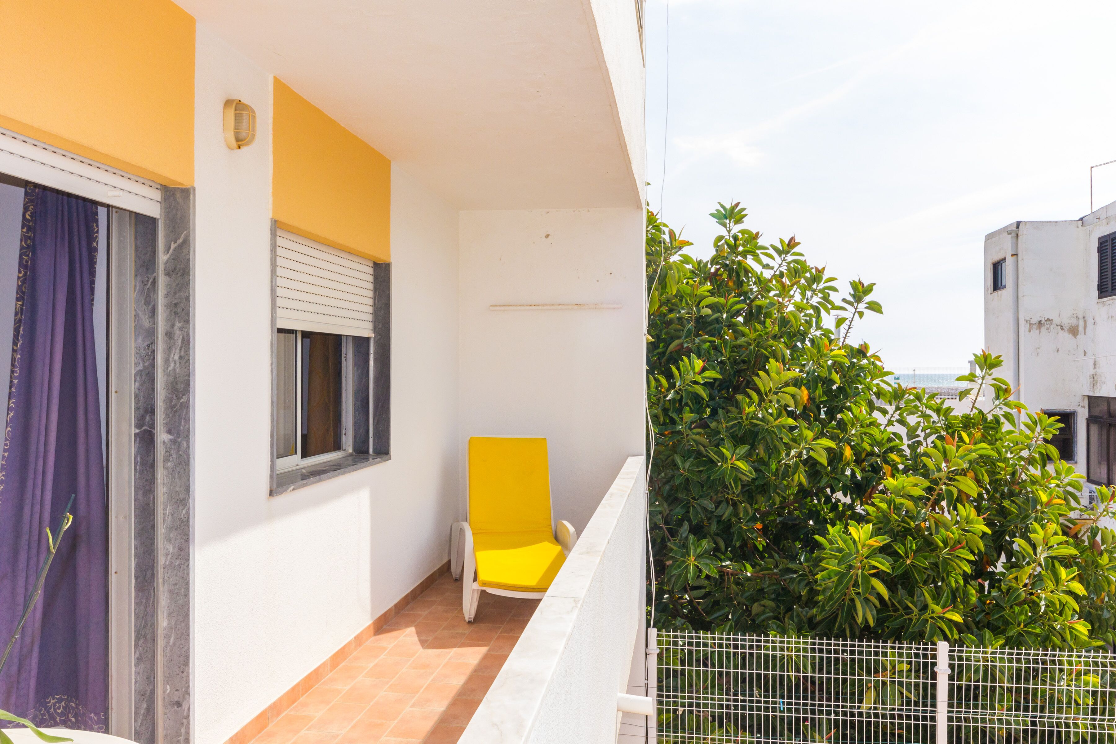 A balcony with a yellow chair, a window with blinds, and a white fence.