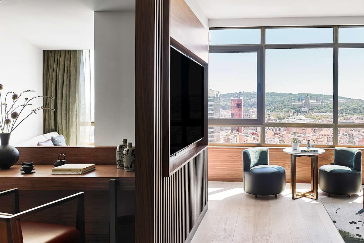Une chambre d’hôtel avec une grande fenêtre, un lit, un bureau avec une télévision, deux chaises et une petite table.