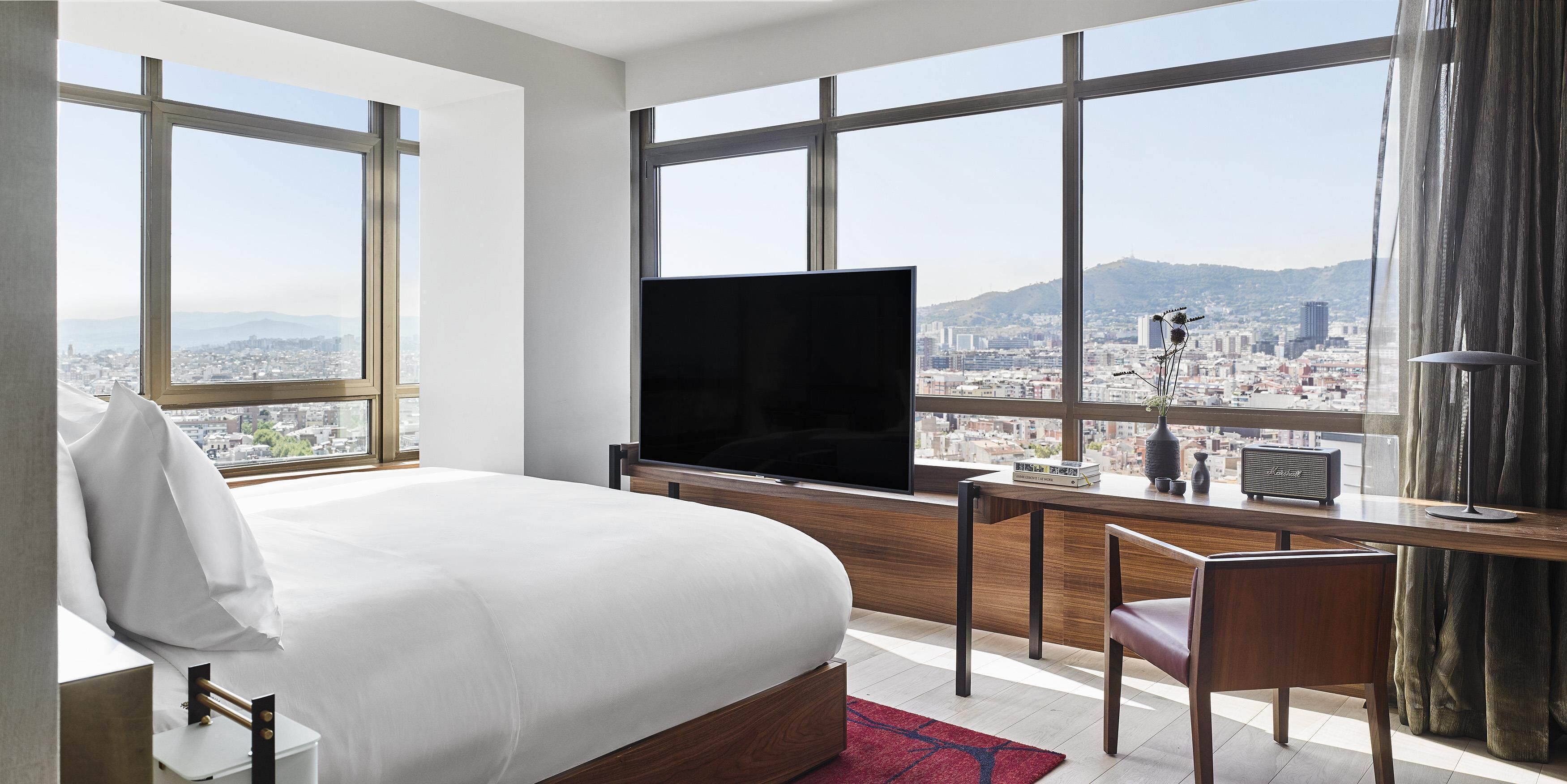 Habitación de hotel con una cama grande, un escritorio con televisor de pantalla plana, una silla y vistas a un paisaje urbano a través de amplias ventanas.