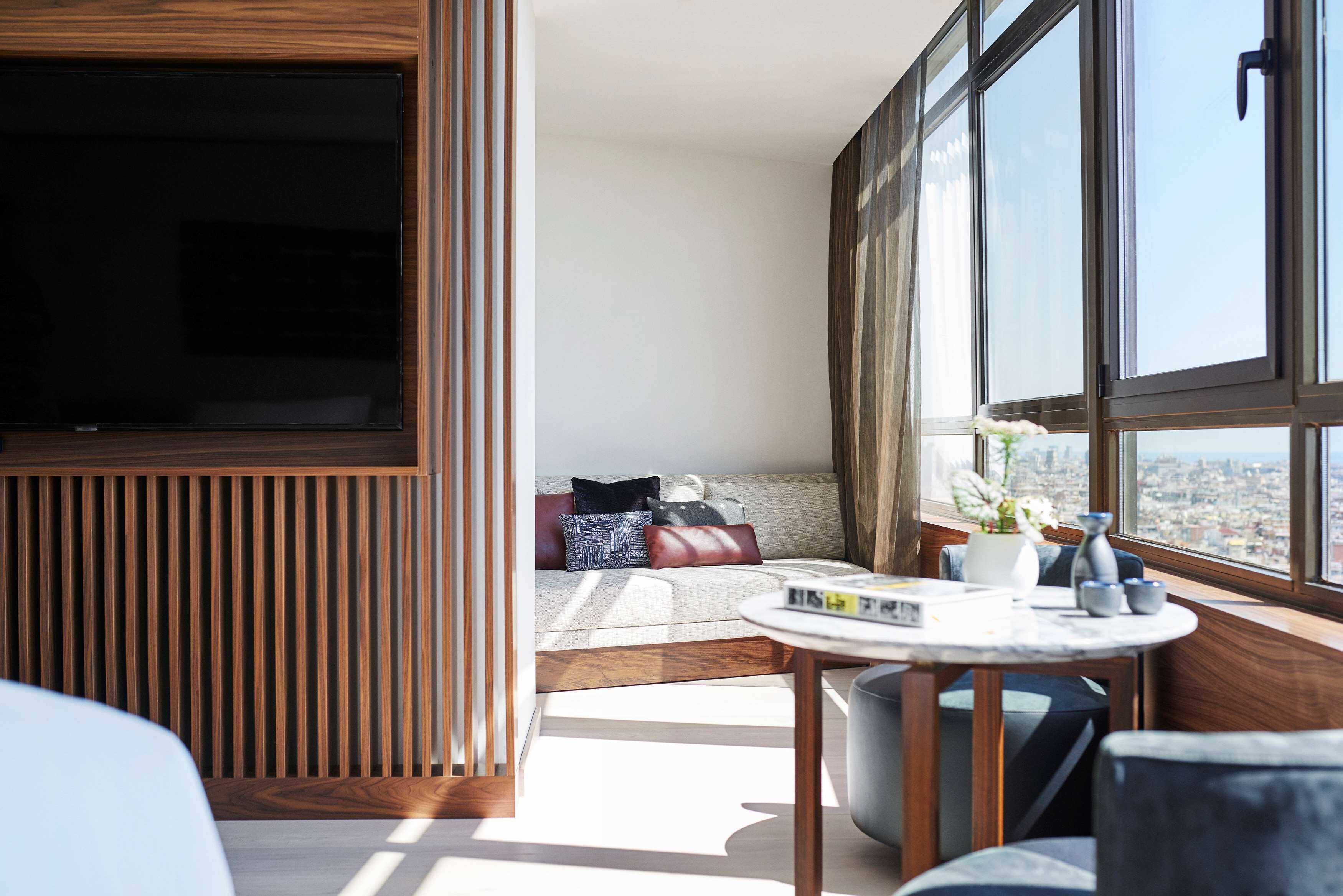 Een moderne woonkamer met een grote televisie, comfortabele zithoek, een klein tafeltje met boeken en een stadszicht door grote ramen.