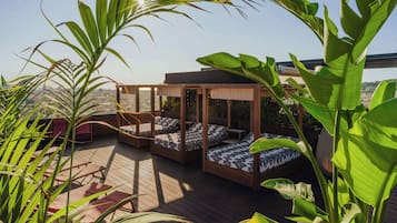 A rooftop deck with wooden flooring, a covered seating area with patterned cushions, and large green plants.