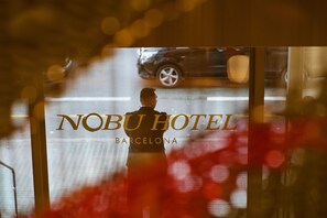 A man in a suit standing in front of a window with a car passing by. The text "Nobu Hotel Barcelona" is visible.