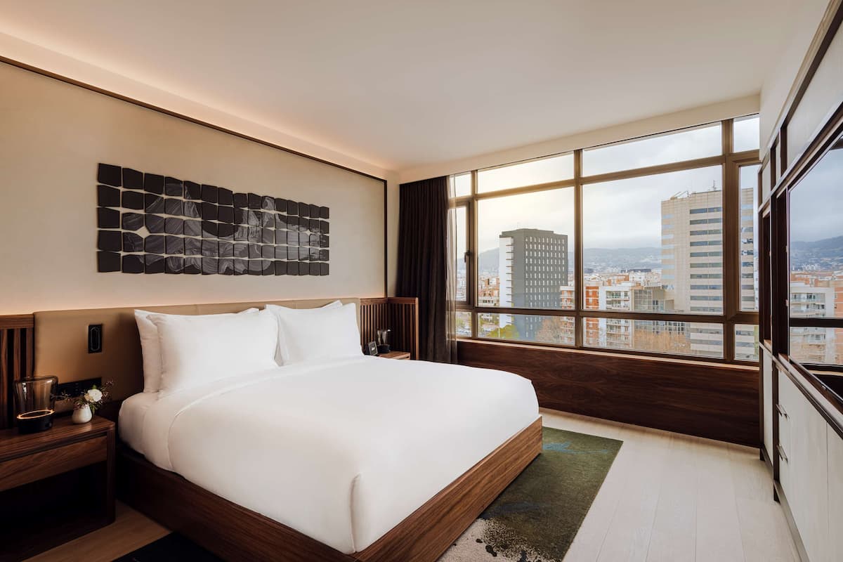 Une chambre d’hôtel moderne dotée d’un grand lit, offrant une vue sur la ville grâce à de grandes fenêtres du sol au plafond, et agrémentée d’un élément décoratif mural.