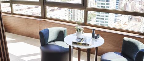 A modern room with two blue armchairs, a small round table with a book and a plant, and a city view through large windows.
