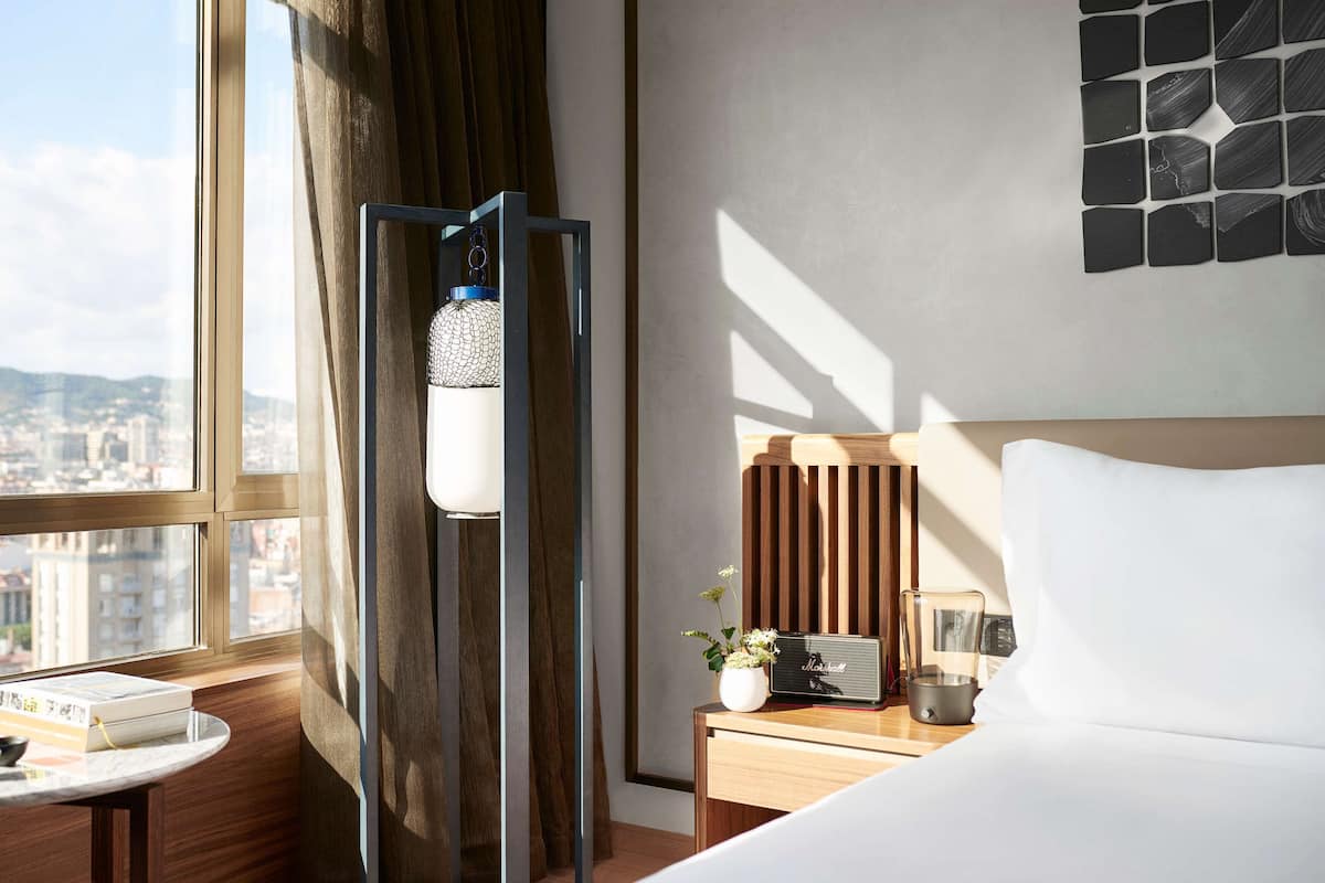 Une chambre d’hôtel avec un lit, une table de chevet avec une radio, une petite table avec des livres, une fenêtre avec des rideaux et une vue sur la ville.