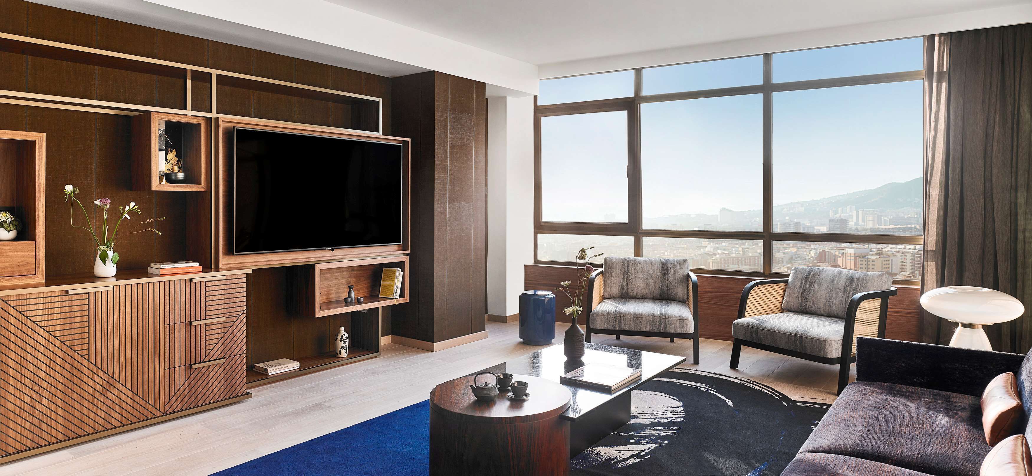 Uma sala de estar moderna com uma televisão grande, assentos confortáveis, uma mesa de centro e vista para a cidade através de grandes janelas.