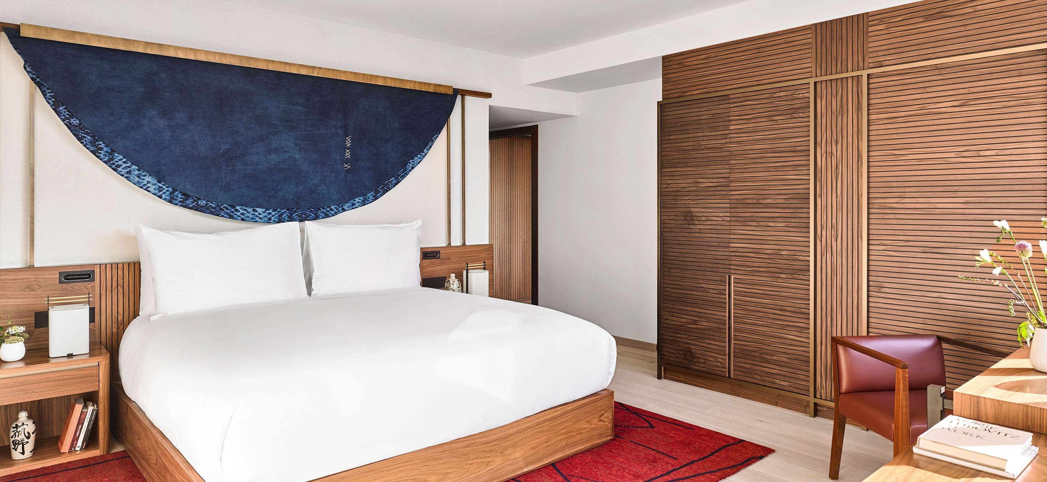 Een moderne slaapkamer met een groot bed, een houten hoofdbord, een nachtkastje met een lamp, een rood vloerkleed en een houten garderobe.
