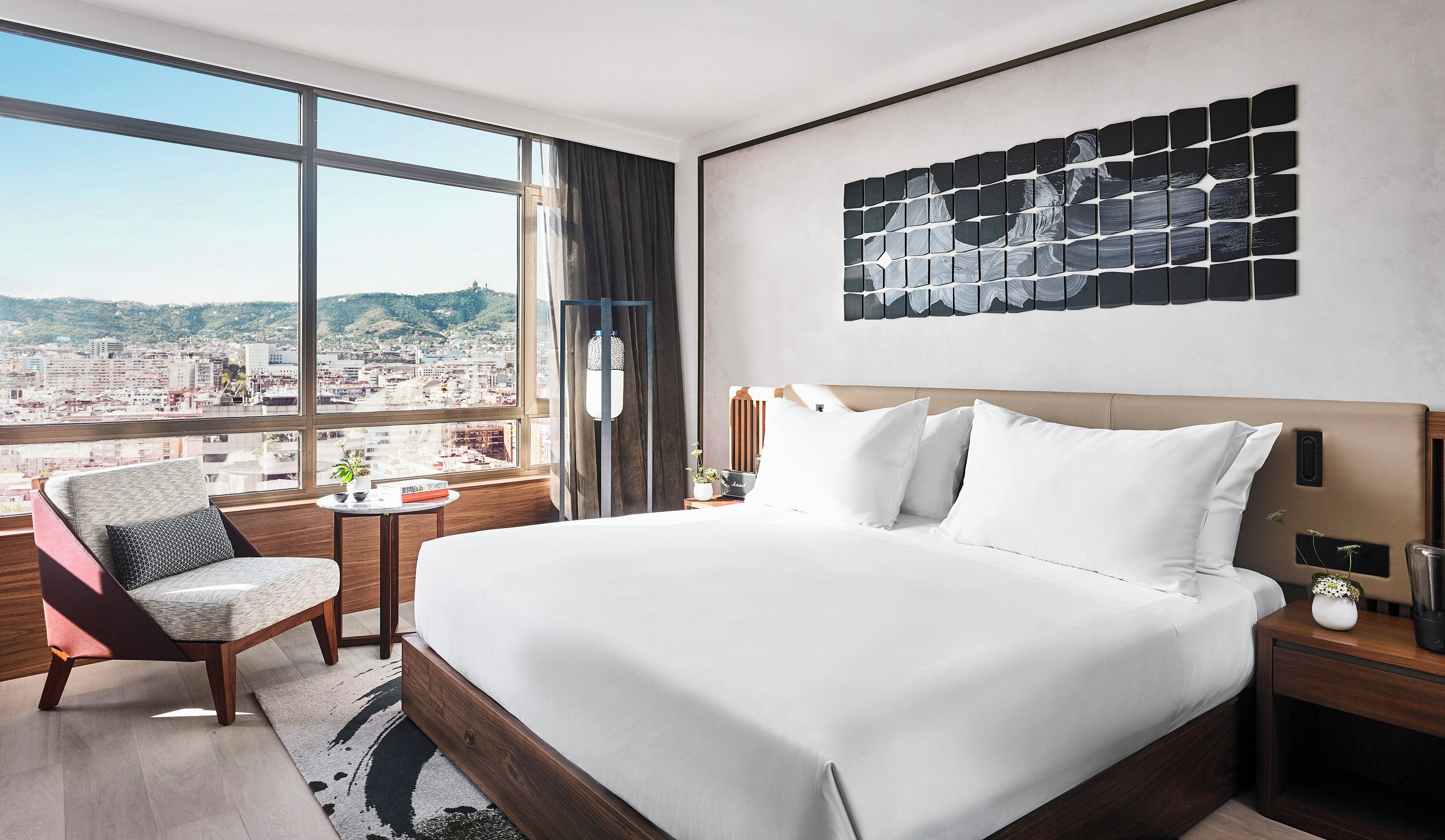 Een moderne hotelkamer met een groot bed, een zithoek met een stoel, een klein tafeltje met een plant en uitzicht op de stad door grote ramen.