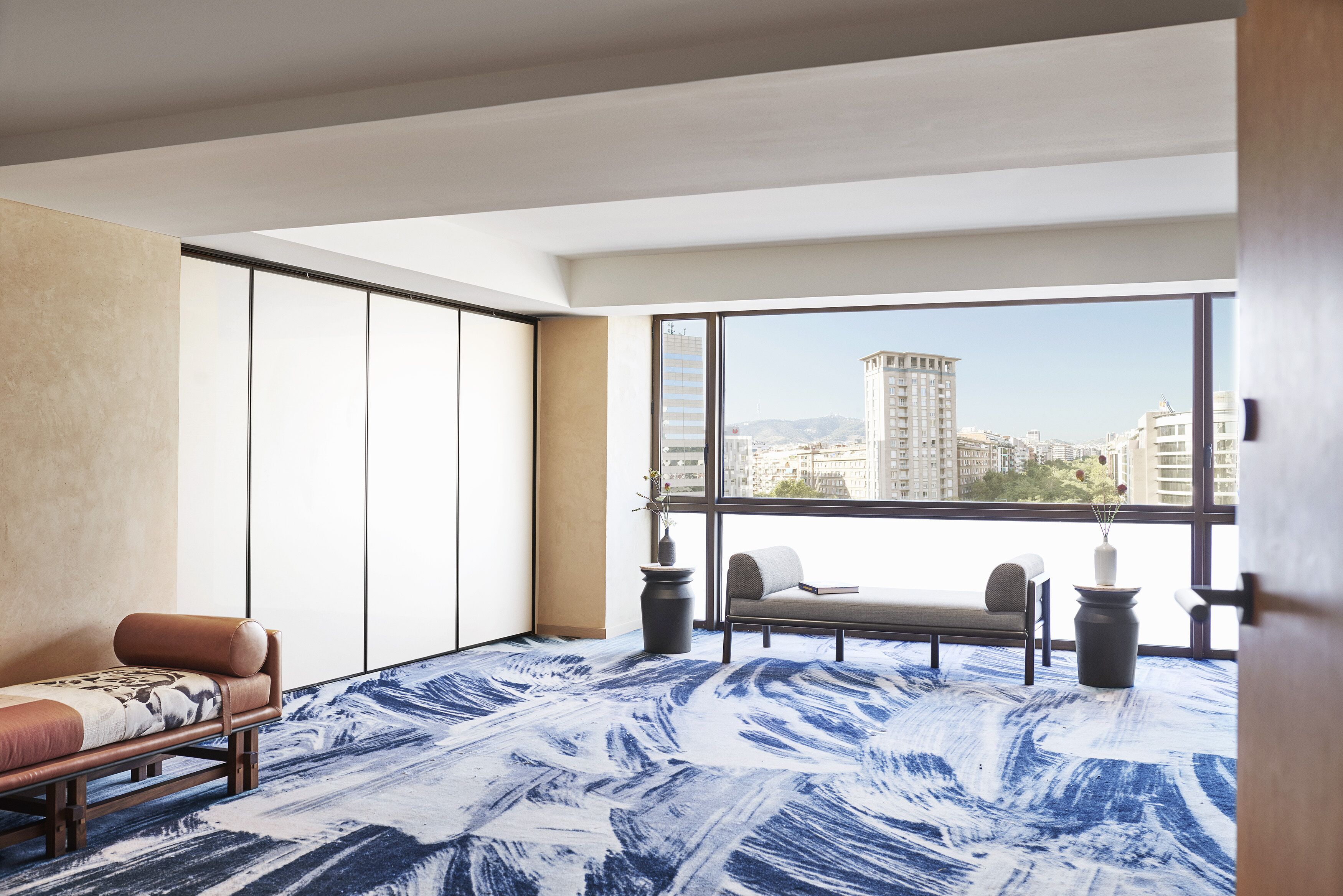 Quarto de hotel moderno com uma janela grande, um sofá, uma cadeira e um tapete estampado.