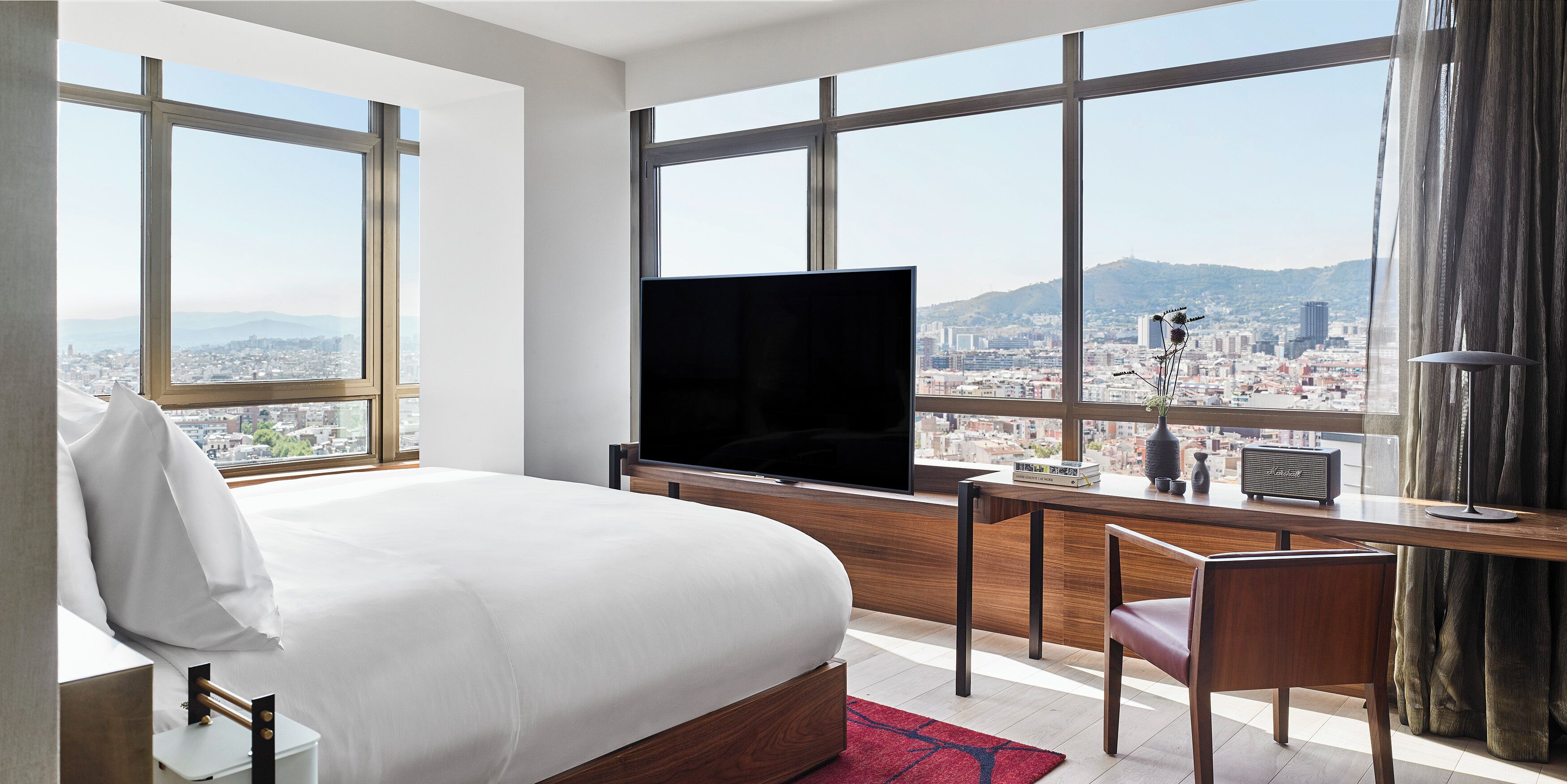 Habitación de hotel con cama, escritorio, silla, televisor, un jarrón con flores y vistas a un paisaje urbano a través de amplias ventanas.