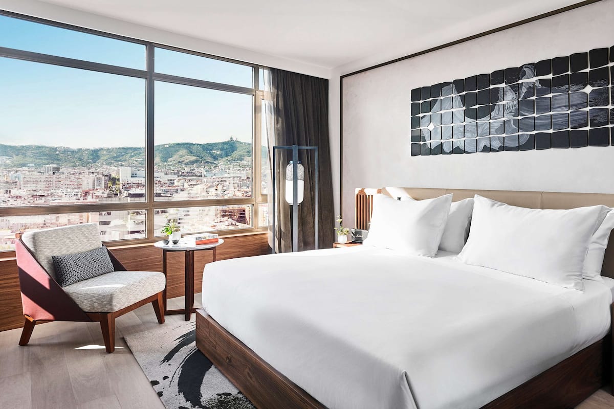 Une chambre d’hôtel moderne, dotée d’un grand lit, d’une chaise, d’une petite table et offrant une vue sur la ville grâce à de grandes fenêtres.