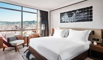 Una habitación de hotel moderna con una cama grande, una silla, una mesita y vistas de la ciudad a través de amplias ventanas.