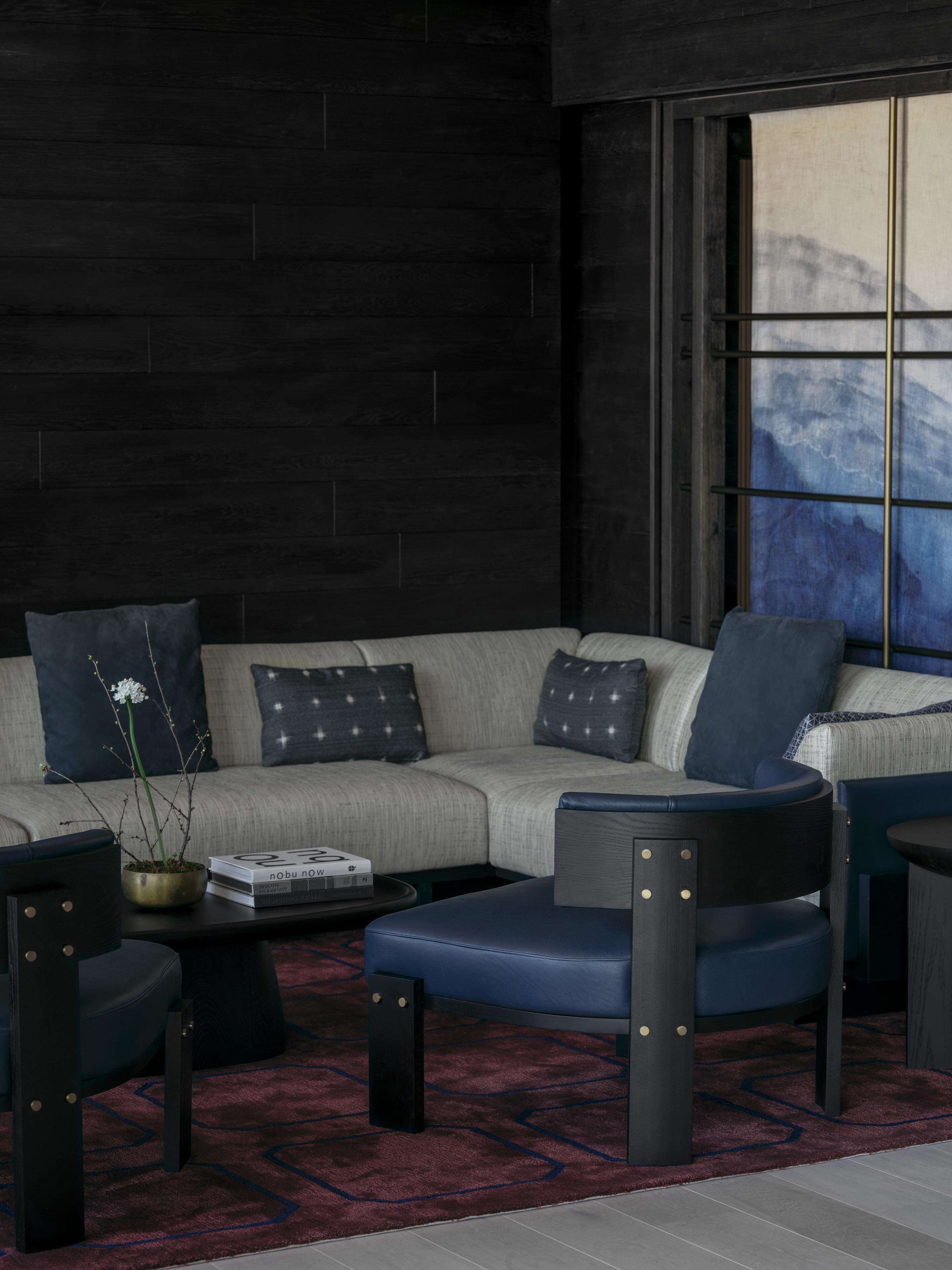 Una sala moderna con una pared de madera oscura, un sofá gris con cojines estampados, un sillón azul, una pequeña mesita de noche con una planta y una ventana con vistas a la montaña.