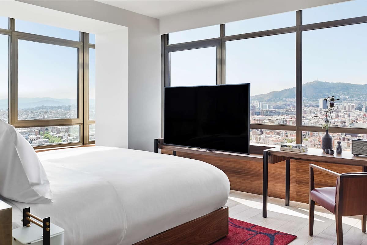 Une chambre d’hôtel avec un grand lit, un bureau avec une télévision et une vue sur le paysage urbain grâce à de grandes fenêtres.