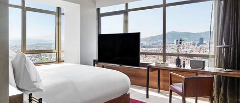 A hotel room with a large bed, a desk with a TV, and a view of a cityscape through large windows.