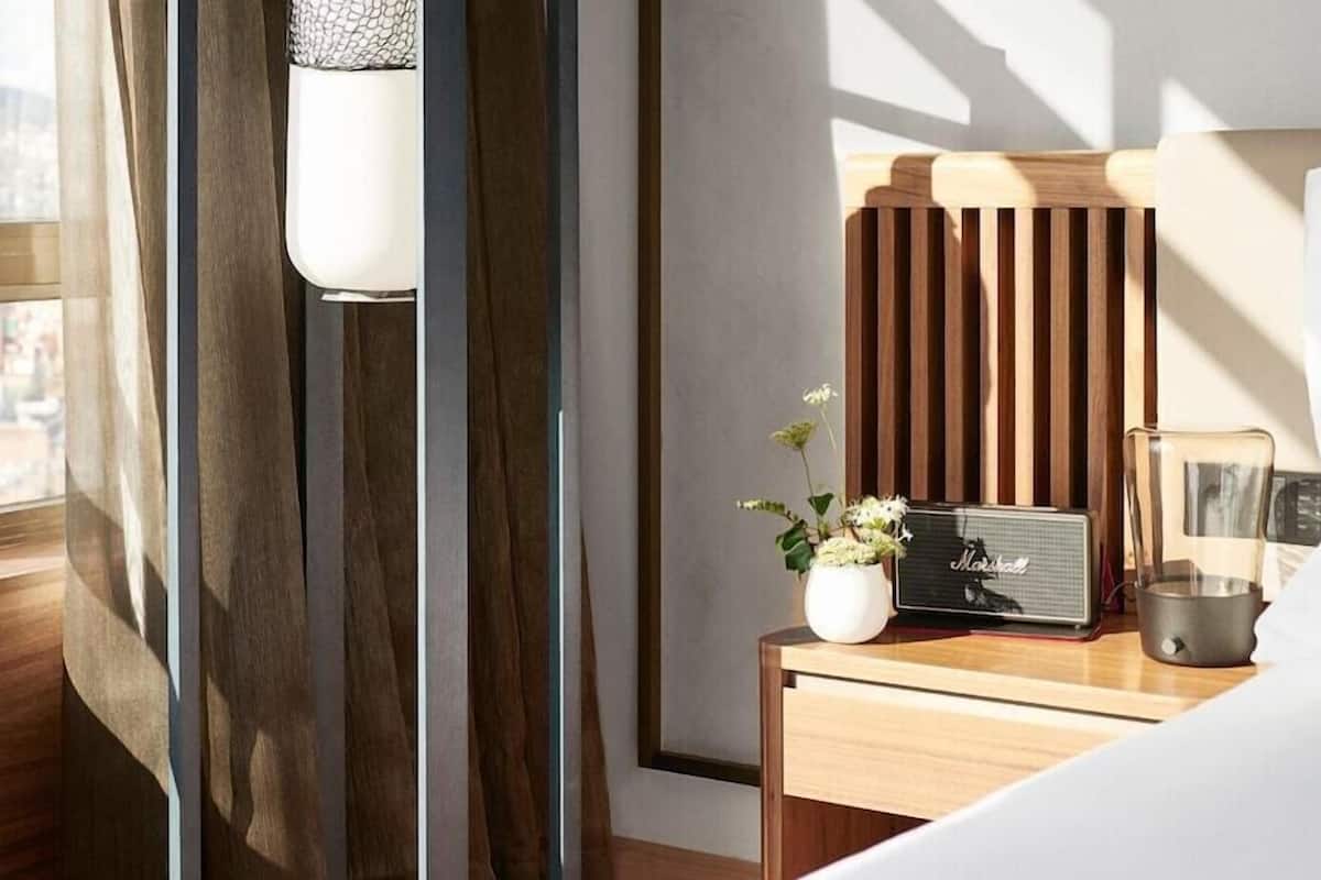 Une chambre d’hôtel avec un lit, une table de chevet avec une radio, un vase de fleurs et une fenêtre avec des rideaux.