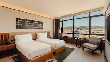Habitación de hotel con dos camas, una silla, una mesita, vista a la ciudad y una obra de arte en la pared.