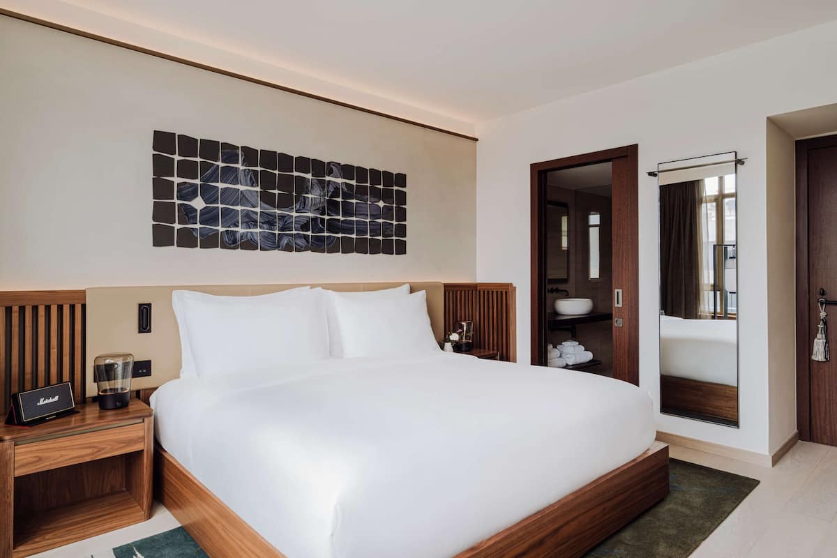 Une chambre d’hôtel moderne avec un grand lit, une tête de lit en bois, une table de chevet avec une radio, une œuvre d’art murale et une salle de bain visible à travers une porte ouverte.
