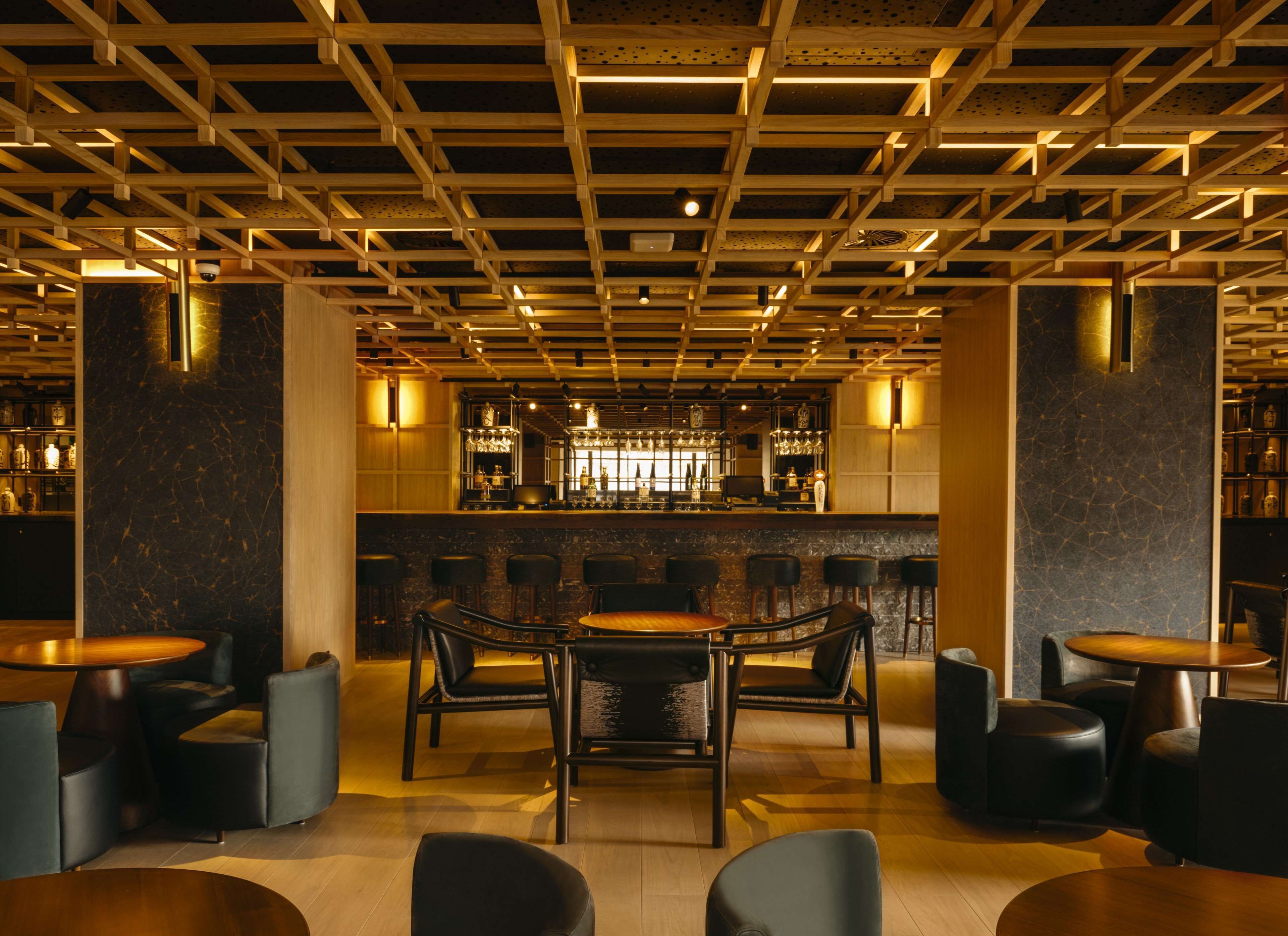 Een bar met houten balken in het plafond, barstoelen en een bargedeelte met flessen.