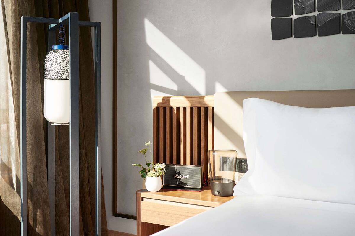 Une chambre d’hôtel moderne avec une tête de lit en bois, une table de chevet dotée d’une radio, une petite plante et une lampe décorative.