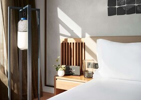 A modern hotel room with a wooden headboard, a bedside table with a radio, a small plant, and a decorative lamp.