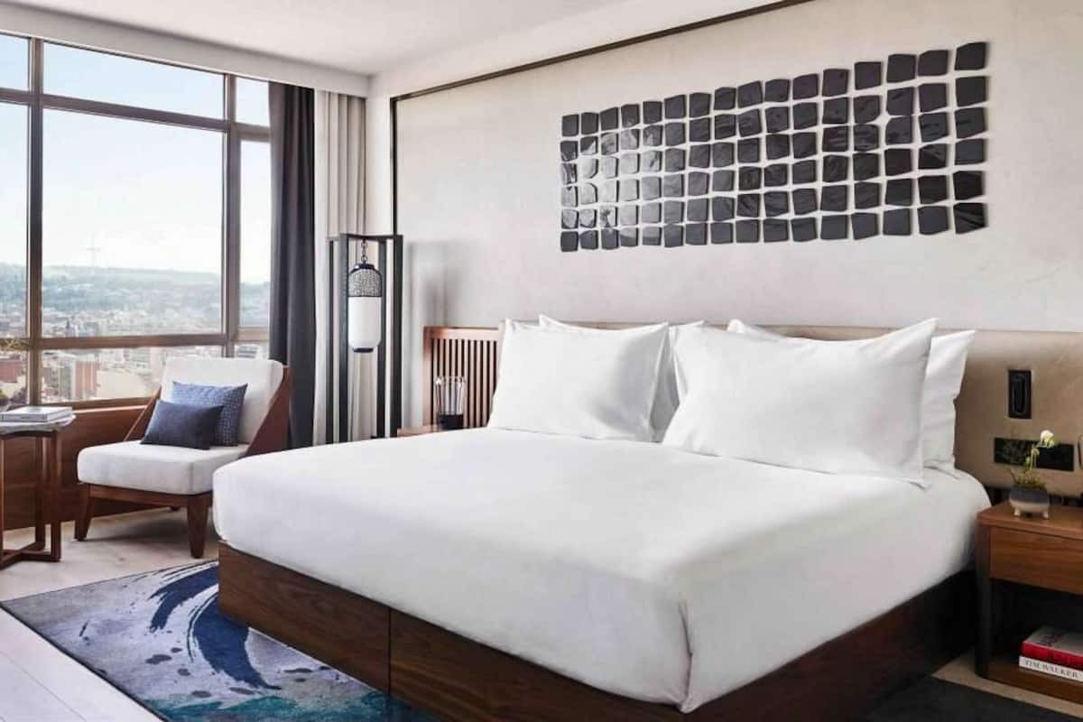Une chambre d’hôtel moderne avec un grand lit, une vue sur la ville grâce à de grandes fenêtres du sol au plafond, une œuvre d’art accrochée au mur et une petite table d’appoint avec un livre.