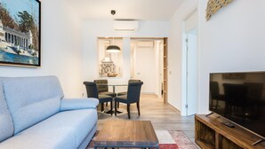 Ein modernes Wohnzimmer mit einer Couch, einem Couchtisch und einem Fernseher auf einem Holzgestell. Es gibt einen Essbereich mit Tisch und Stühlen, und einen Flur, der zu einer Tür führt.