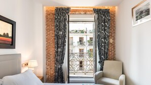 Ein Hotelzimmer mit Bett, Stuhl, einem Balkon mit schmiedeeisernem Geländer und Blick auf die Stadt.