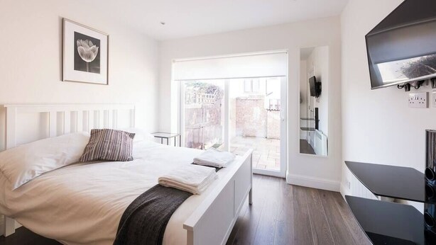 A modern bedroom with a large bed, a TV mounted on the wall, and a sliding glass door leading to an outdoor area. - Spectacular Modern Apartment (5 mins to tube) (Greater London)