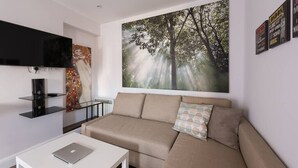 A modern living room with a beige sofa, a flat-screen TV mounted on the wall, and a large forest-themed painting above the sofa. - Spectacular Modern Apartment (5 mins to tube) (Greater London)